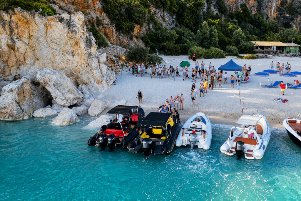 Crowded beach with a rocky cliff, trees, and many boats anchored along the shore on turquoise water.